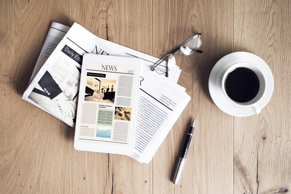 A newspaper, a tablet, a pair of glasses, a pen, and a cup of coffee rest on a table.