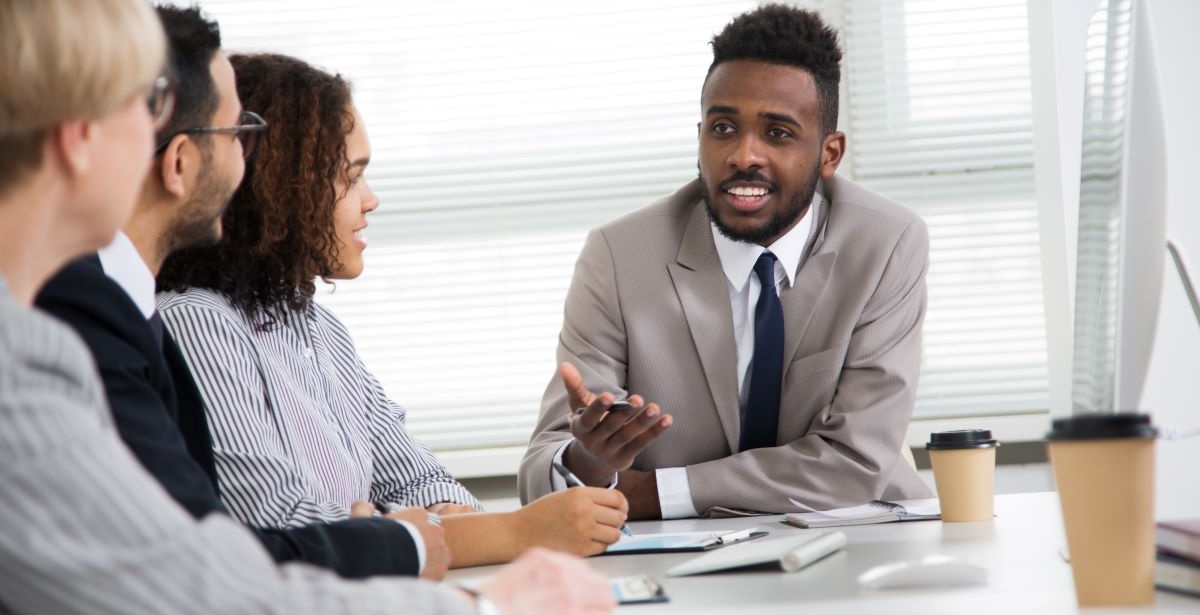 A businessperson addresses colleagues gathered around a conference table.